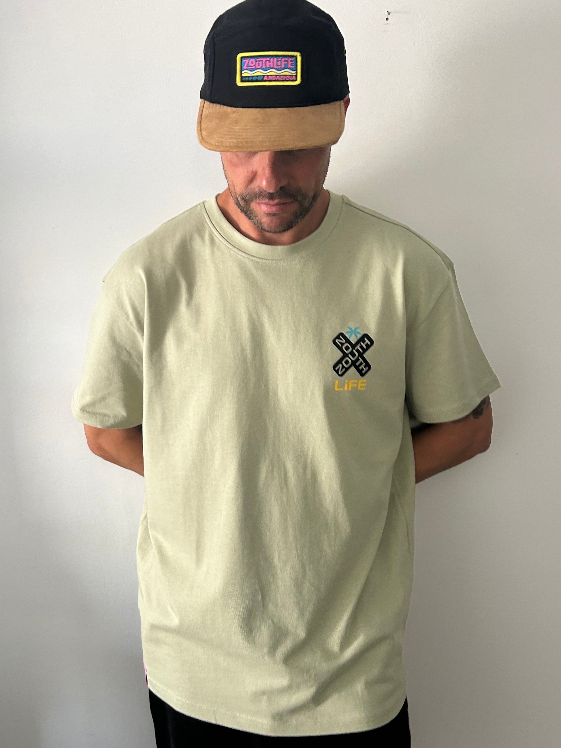 Vista frontal de hombre vistiendo la camiseta oversize Shokito en color verde oliva. La prenda  presenta un pequeño logo en forma de "X" negra con el texto Zouth Life en el lado derecho del pecho. El hombre también lleva una gorra de dos tonos (corona negra y visera de color canela) con un parche rectangular colorido de Zouth Life. 