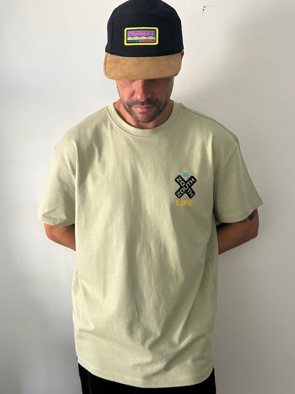 Vista frontal de hombre vistiendo la camiseta oversize Shokito en color verde oliva. La prenda  presenta un pequeño logo en forma de "X" negra con el texto Zouth Life en el lado derecho del pecho. El hombre también lleva una gorra de dos tonos (corona negra y visera de color canela) con un parche rectangular colorido de Zouth Life. 