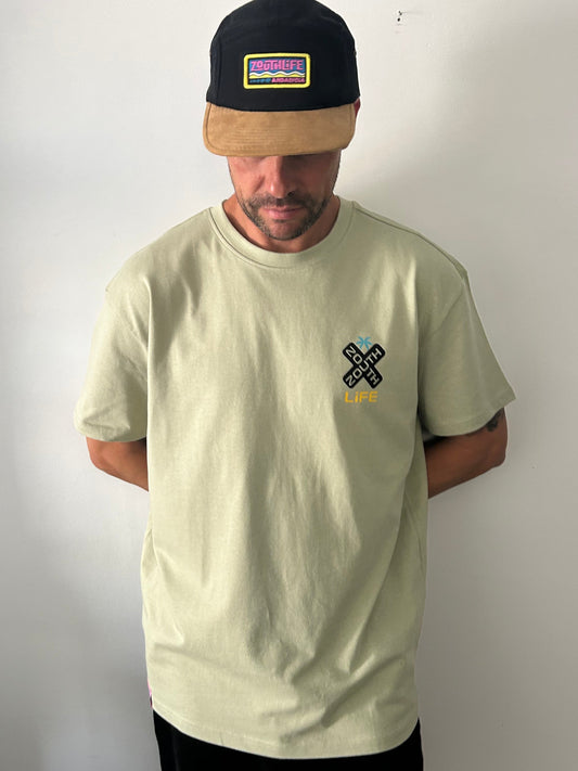 Vista frontal de hombre vistiendo la camiseta oversize Shokito en color verde oliva. La prenda  presenta un pequeño logo en forma de "X" negra con el texto Zouth Life en el lado derecho del pecho. El hombre también lleva una gorra de dos tonos (corona negra y visera de color canela) con un parche rectangular colorido de Zouth Life. 