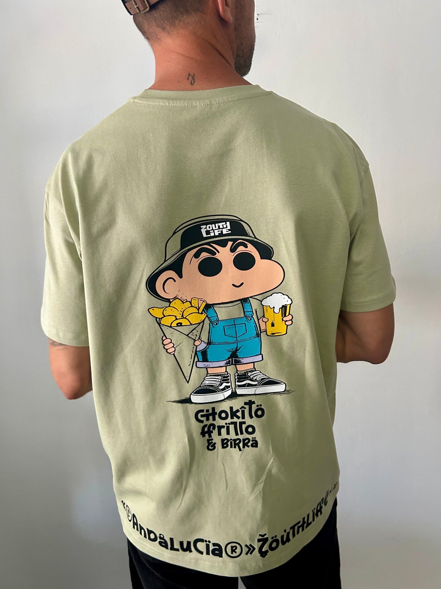 parte trasera de la Camiseta Oversize Shokito en color verde oliva. El diseño presenta una ilustración estilo anime del personaje Shin-chan vistiendo ropa urbana con un gorro, sosteniendo un cucurucho de chocos fritos en una mano y una jarra de cerveza en la otra. Debajo del personaje se lee el texto 'Chokito frito & birra', y en el dobladillo inferior de la camiseta destaca la frase 'Andalucía Zouth Life' 