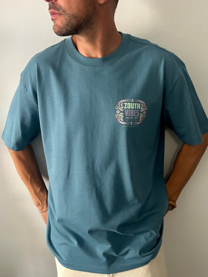Vista frontal de un hombre modelando la camiseta oversize Tribal Vibes de Zouth Life en color azul petróleo. La prenda cuenta con un logotipo circular pequeño en el lado izquierdo del pecho que replica el diseño tribal y playero de la espalda en tonos pastel.