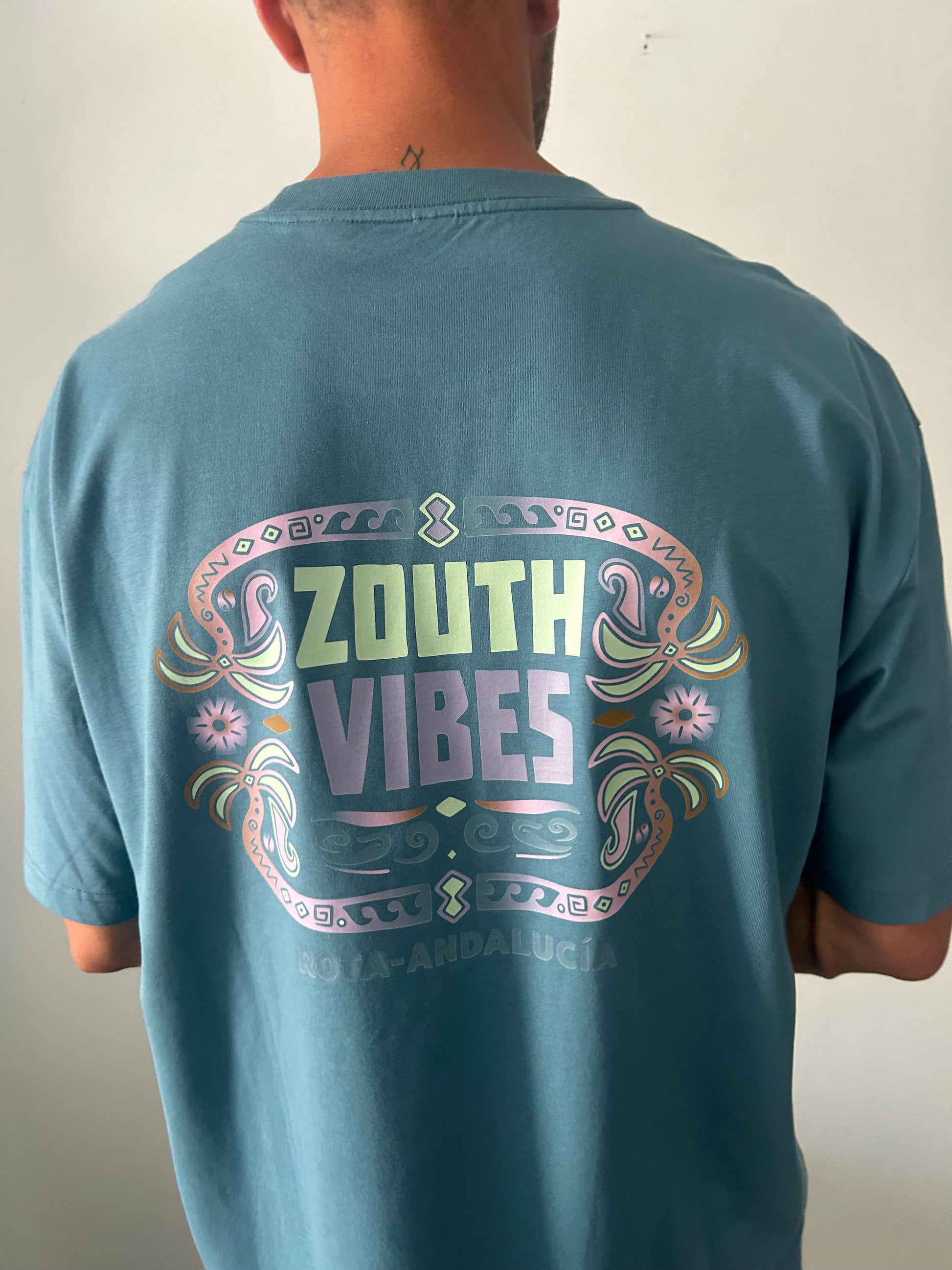 Vista trasera de un hombre modelando la camiseta oversize Tribal Vibes de Zouth Life en color azul petroleo.El diseño de la espalda destaca por una gran gráfica de estilo tribal y surfero en tonos pastel verdes, lilas y rosados, donde se lee 'ZOUTH VIBES' enmarcado por palmeras, olas y patrones geométricos.