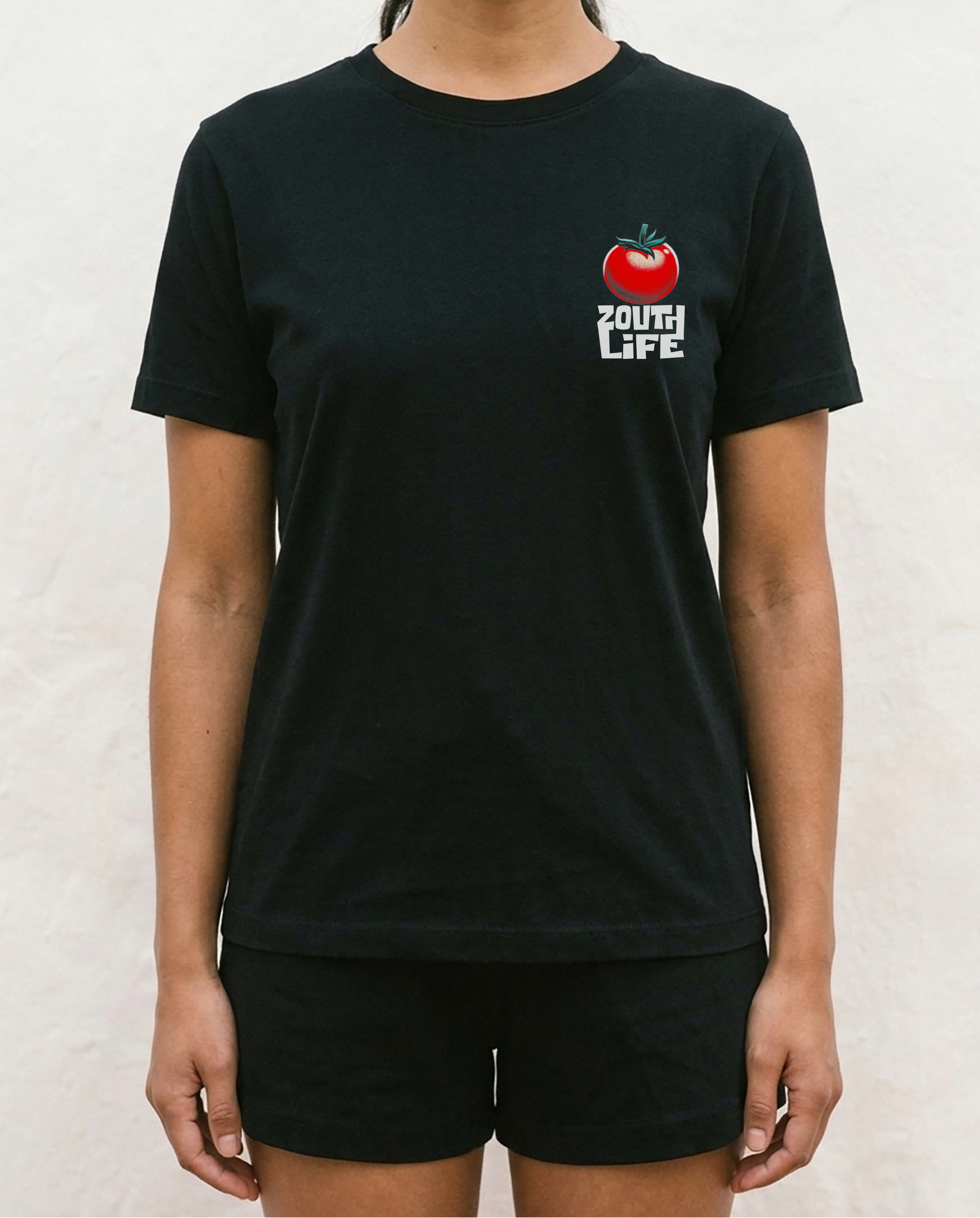 Vista frontal de primer plano de la camiseta negra regular fit Blanca Nieves de la marca Zouth Life. La prenda presenta un diseño minimalista en el lado izquierdo del pecho con la ilustración de un tomate rojo brillante y el logotipo de la marca 'ZOUTH LIFE' en letras blancas justo debajo, modelada frente a una pared blanca.