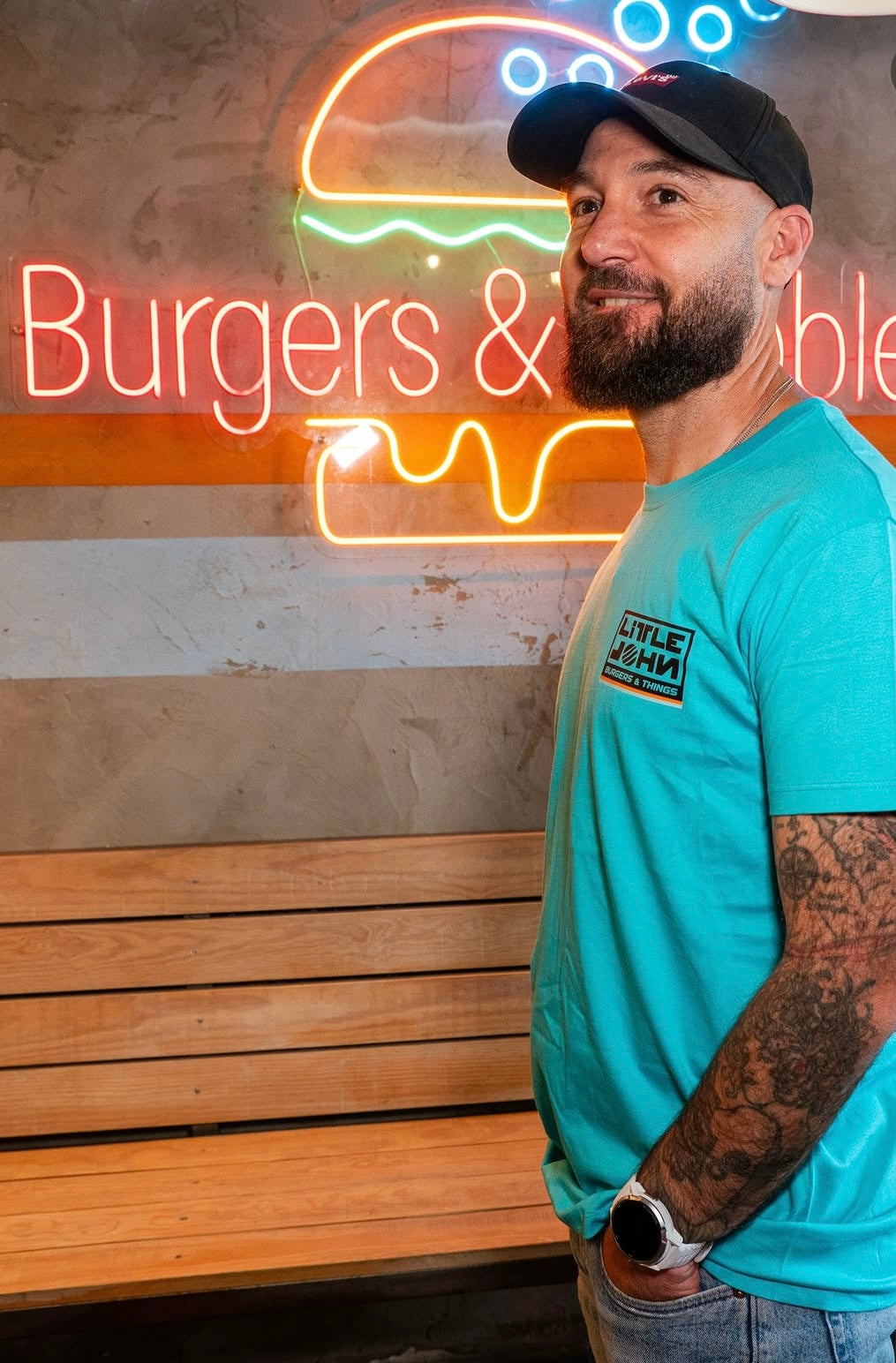 Camiseta azul turquesa Little Burger Retro de Zouthlife. Foto lifestyle de modelo con gorra negra y tatuajes posando junto a un neón de hamburguesería. Detalle del logo cuadrado en el lateral del pecho.