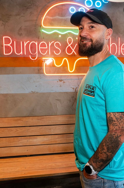 Camiseta azul turquesa Little Burger Retro de Zouthlife. Foto lifestyle de modelo con gorra negra y tatuajes posando junto a un neón de hamburguesería. Detalle del logo cuadrado en el lateral del pecho.