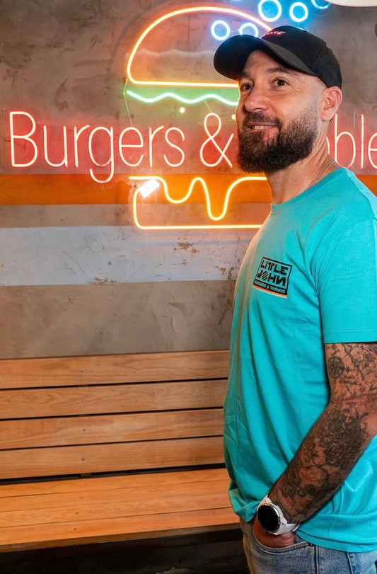 Camiseta azul turquesa Little Burger Retro de Zouthlife. Foto lifestyle de modelo con gorra negra y tatuajes posando junto a un neón de hamburguesería. Detalle del logo cuadrado en el lateral del pecho.