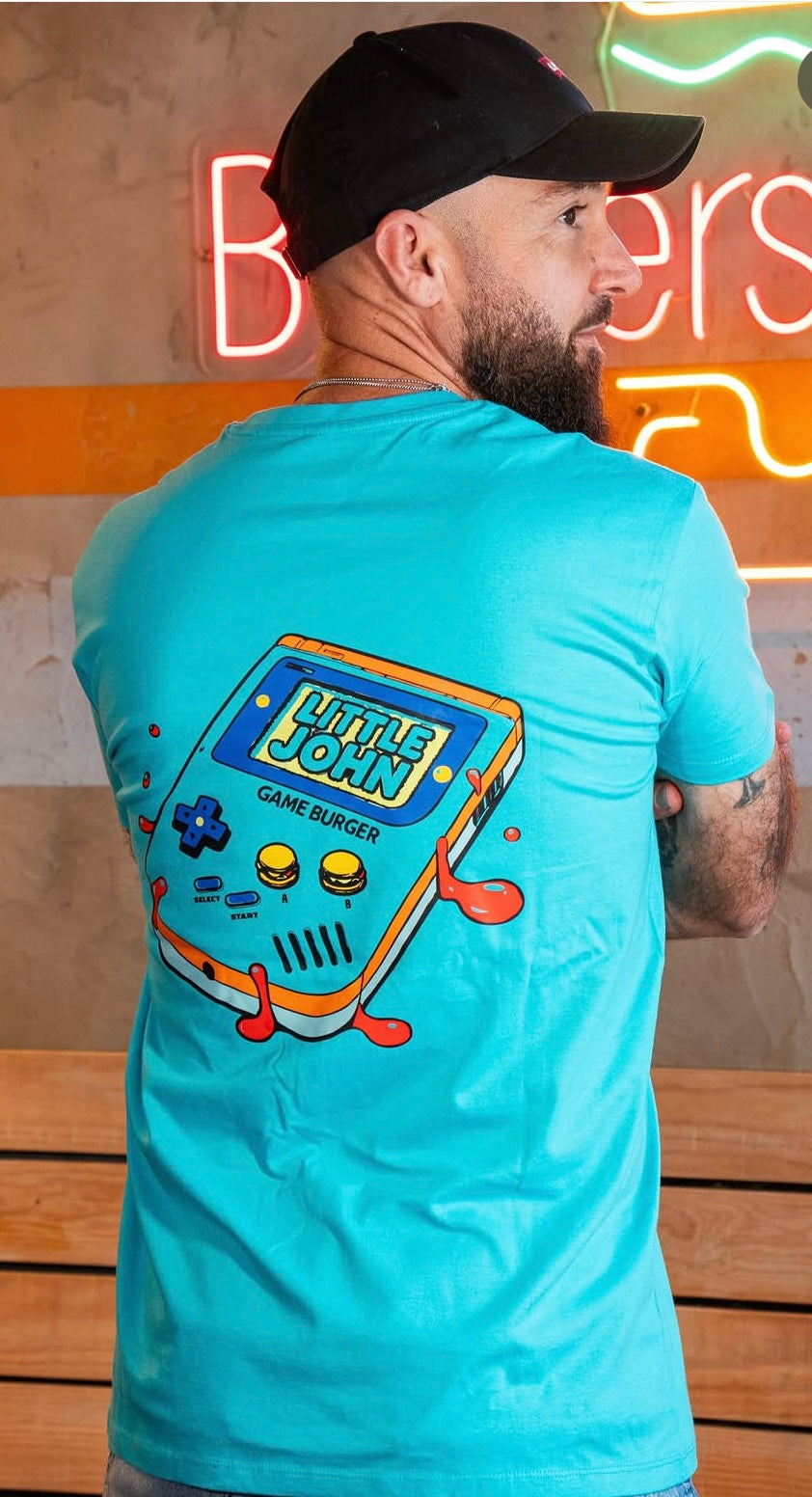Vista posterior de la camiseta azul turquesa de Zouthlife con un diseño gráfico de una consola portátil clásica (tipo Game Boy) en la espalda. La pantalla muestra el texto 'Little John' y debajo 'Game Burger', con botones en forma de mini hamburguesas y detalles de pintura roja chorreando.