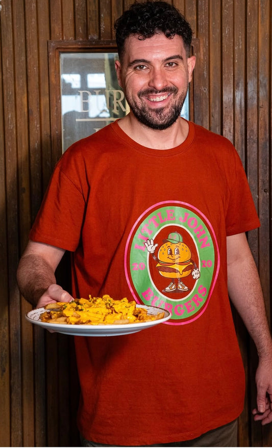 Hombre sonriente con camiseta Little Burger Retro de color terracota, sosteniendo un plato de patatas fritas con queso cheddar. El diseño central muestra el logo circular de 'Little John Burgers 2020' con una ilustración de hamburguesa animada sobre fondo verde y rosa.