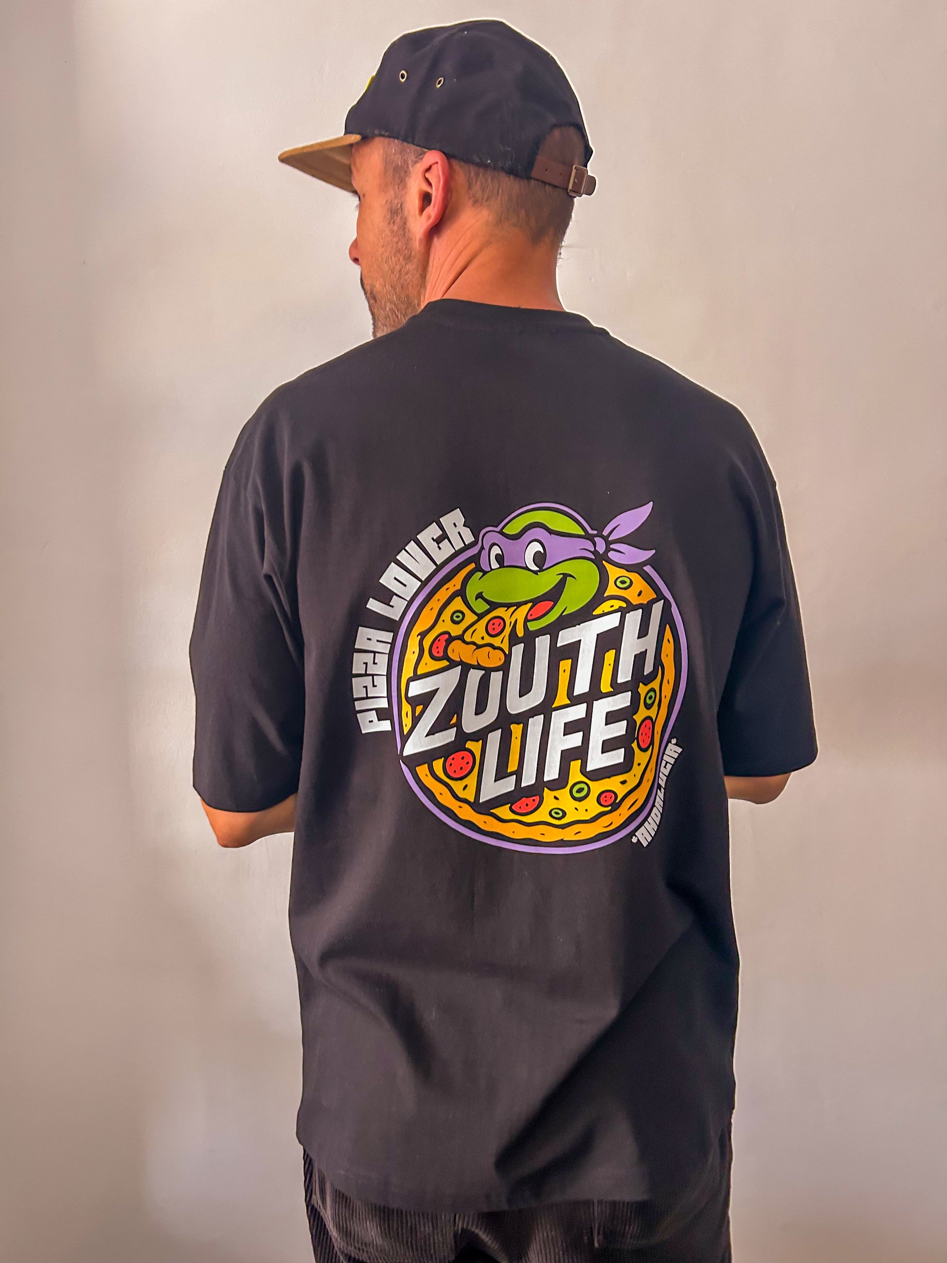 Primer plano de la parte trasera de una camiseta negra de corte oversize de la marca Zouth Life. Destaca un gráfico circular grande de estilo retro con una tortuga ninja comiendo una porción de pizza, rodeada por el texto "Pizza Lover" y el logotipo de la marca. El modelo lleva una gorra negra con visera marrón y pantalones de pana oscuros.