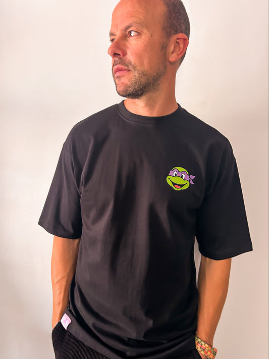 Vista frontal de un hombre vistiendo la camiseta negra de algodón 240 gramaje "Pizza Lover" de Zouth Life. La prenda presenta un estampado  minimalista en el pecho izquierdo con la cara de una tortuga ninja con antifaz morado. El modelo posa de perfil con las manos en los bolsillos, mostrando el ajuste holgado de las mangas y el cuello redondo.