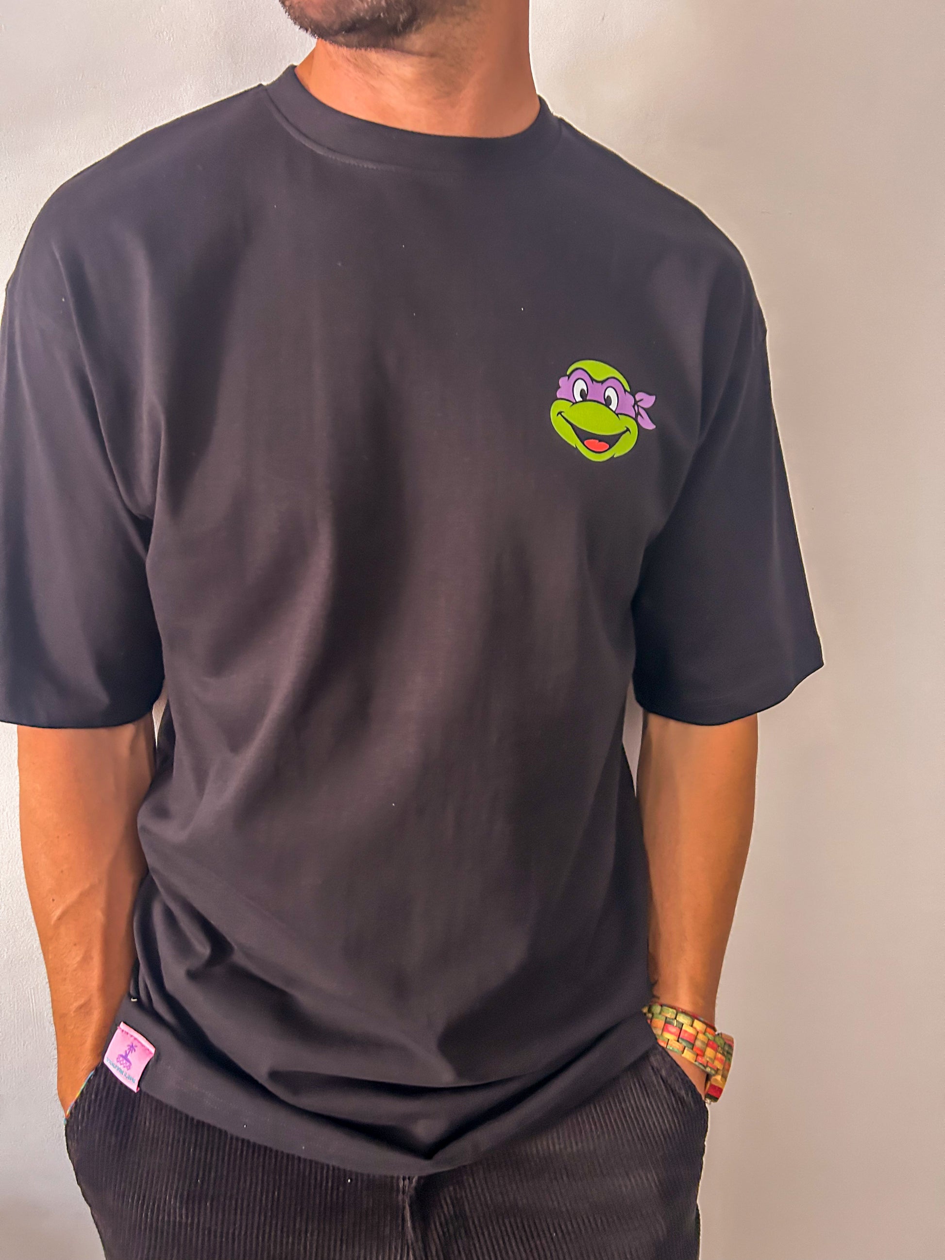 Vista frontal de un hombre vistiendo la camiseta negra de algodón 240 gramaje "Pizza Lover" de Zouth Life. La prenda presenta un estampado  minimalista en el pecho izquierdo con la cara de una tortuga ninja con antifaz morado. El modelo posa de perfil con las manos en los bolsillos, mostrando el ajuste holgado de las mangas y el cuello redondo.